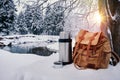 Backpack of traveller, thermos and enameled mug of coffee or tea against snowy winter landscape background