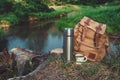 Backpack of traveller, thermos and Enameled mug of coffee or tea on bank of beautiful forest river