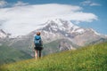 Backpacker woman with a backpack walking and enjoying snowy slopes of Kazbek 5054m mountain while she walking by green grass hill Royalty Free Stock Photo