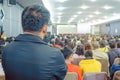 Backside view of spectators standing in a gathering in the back of auditorium full of people.