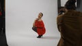 Backstage of model and professional team in the studio. Woman model posing in red dress sitting on knees, photographer Royalty Free Stock Photo