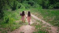 Backwards view of little identical twin sisters with long hair walking together holding hands at road among field Royalty Free Stock Photo