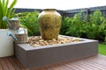 Backyard setting unfolds with large decorative vase, two watering cans, wooden deck, green trees, and wooden fence backdrop