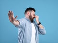 Bad smell. Young bearded guy closing his nose and showing STOP gesture, expressing disgust on blue studio background