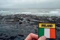 Badge with sign Ireland and Irish National flag in focus. Stone bed of the ocean and surfer with blue board out of focus. Lahinch