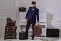 Baggage delivery concept. Macho elegant on strict face stands near pile of vintage suitcase, holds suitcase. Man