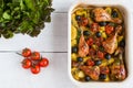 Baked chicken drumsticks in red dish. Cooked with cherry tomatoes, black olives, rosemary and potatoes. White wooden table