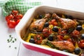 Baked chicken drumsticks in red dish. Cooked with cherry tomatoes, black olives, rosemary and potatoes. White wooden table Royalty Free Stock Photo