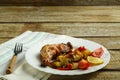 Baked chicken leg with potatoes and tomato on a white plate with a fork on a napkin on a wooden table. Royalty Free Stock Photo