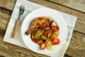 Baked chicken leg with potatoes and tomato on a white plate with a fork on a napkin on a wooden table. Royalty Free Stock Photo