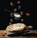 Baked round pie with plums on a levitating wooden kitchen board, top of a sieve with powdered sugar and livitating slices of plum