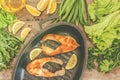 Baked steak trout fish in cast-iron form, around greens, leaves, lettuce, lemon, olive oil, pepper, condiments. From above, close