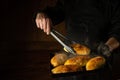 The baker bakes hot dog buns. Tongs in chef hand and baking sheet with hot pies. Place for advertising on a dark background