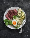 Balanced lunch - medium roast beef steak, grilled zucchini and couscous, avocado, spinach, egg salad on a dark background, top