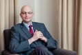 Bald attractive businessman in grey suit, blue shirt, red tie and glasses sitting in a chair in a hotel room. Royalty Free Stock Photo