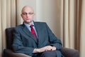 Bald attractive businessman in grey suit, blue shirt, red tie and glasses sitting in a chair in a hotel room Royalty Free Stock Photo