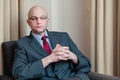 Bald attractive businessman in grey suit, blue shirt, red tie and glasses sitting in a chair in a hotel room Royalty Free Stock Photo