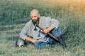 Bald bearded man in linen shirt sits in field, holding an ax, leaning on his helmet. Calm look. Blurred background