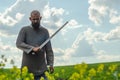 Bald bearded man in metal chain mail over linen shirt stands in the middle of field, holding a sword. Militant look. Blurred