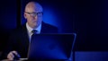 Bald businessman, in a jacket with a tie and glasses, middle-aged, sitting in the dark behind a laptop, looking at the Royalty Free Stock Photo