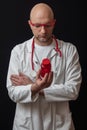 Bald doctor in white coat and red glasses with red stethoscope reading information on a red bottle with pills. Black background,