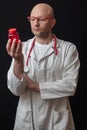 Bald doctor in white coat and red glasses with red stethoscope reading information on a red bottle with pills. Black background,