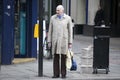 A bald, elderly man in a gray cloak stands at a traffic light, waiting to cross the street