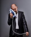 Bald fun tired unhappy loser failed business man punishing himself holding the home iron near the face in suit on grey background