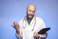 Bald hairless doctor on blue background. Male in his 40s with grey beard. Holding black file and looking at thermometer. Dramatic Royalty Free Stock Photo