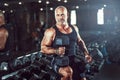 Bald male bodybuilder holds dumbbells in arms while posing in gym in dark. Concept of bodybuilding.