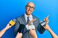 Bald man with beard being interviewed by reporters holding microphones smiling and looking at the camera pointing with two hands