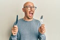 Bald man with beard holding fork and knife ready to eat angry and mad screaming frustrated and furious, shouting with anger