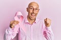 Bald man with beard holding pink cancer ribbon screaming proud, celebrating victory and success very excited with raised arms Royalty Free Stock Photo