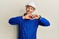 Bald man with beard wearing builder jumpsuit uniform and hardhat smiling in love showing heart symbol and shape with hands