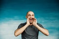 A bald man in a gray T-shirt screams, opening his eyes wide and raising his hands to his mouth like a shout. On a blue background Royalty Free Stock Photo