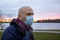 A bald man in a medical mask against the background of the setting sun and a pond. Concept: a violator of the quarantine Royalty Free Stock Photo