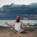 A bald man with a red beard practices yoga on the beach at sunset. A funny dude in a T-shirt and sunglasses meditates on Royalty Free Stock Photo