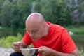 Bald man, soldier in red t-shirt eats from tin bowl outdoor against forest landscape