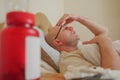 Bald unshaven man with glasses in his bed in focus, Red bottle with pills and thermometer in foreground out of focus, concept flue