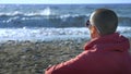 Bald woman in a sweatshirt sits on the seashore during a storm.