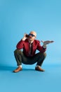 Balded man with mustache wear in vintage style squatting and talking on retro phone looking away over blue background.