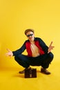 Balded man in tracksuit squats and spreads his arms and looking at camera in glasses over yellow background.