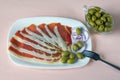 Balkan cuisine . White plate with slices of prsut and green olives on pink pastel background