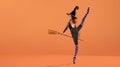 Ballerina on pointe shoes in a black witch costume in a hat and with a broom is dancing on the roof on a Orange background