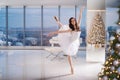 A ballerina in a white tutu poses in a bright hall with a Christmas tree