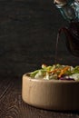 Balsamic vinegar pours from glass jug into a salad in a wooden d