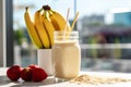 banana and oatmeal smoothie in a glass bottle jar and bananas strawberries on a white kitchen table, sunny blurred outside