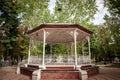 Bandstand in a city park in summer, surrounded by gardens and tall trees, with its typical round stage and metal shelter.