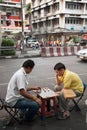 Two men enjoy playing checkers with bottle caps sitting on a noisy street in Bangkok during the day.