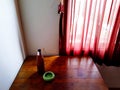 Bottle of beer and green ceramic astray putting on wooden table with red curtain and sunlight from window in the morning.
